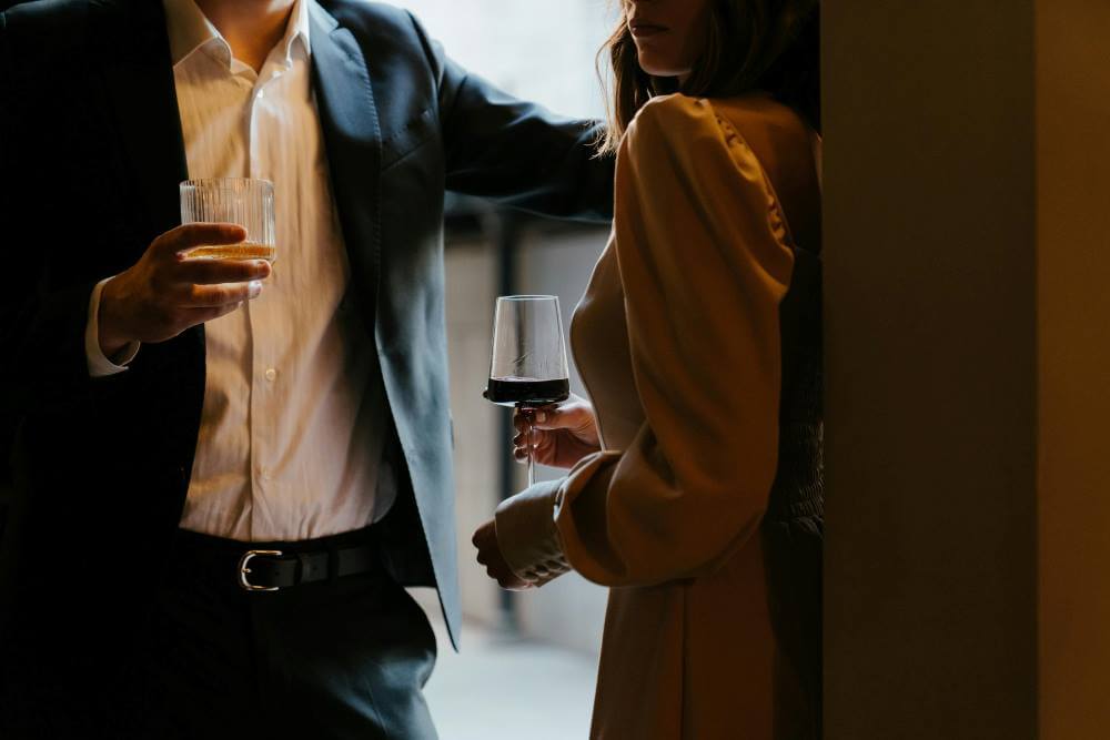 A well-dressed couple holding drinks in a moody setting.